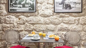 A dining area with a stone wall, two wicker chairs with red cushions, a table with breakfast items, and two framed pictures. - Hotel De Venise (Paris)