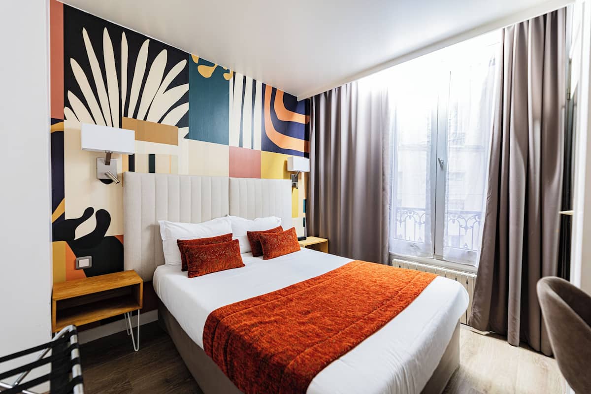 A hotel room with a large bed, a nightstand, a chair, a window with curtains, and a colorful abstract wall design.