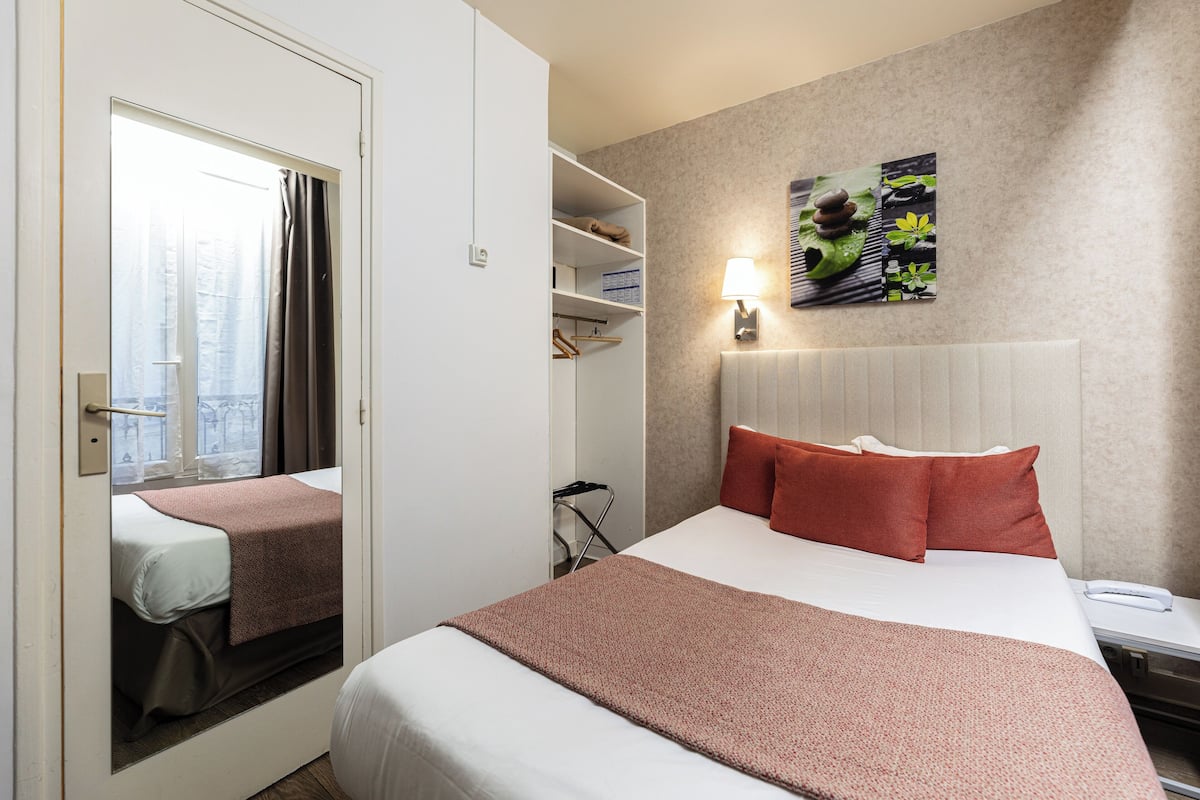 A hotel room with a bed, a bedside table, a lamp, a mirror, a wardrobe, and a shelf with toiletries.