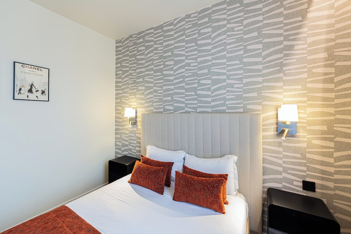 A hotel room with a bed, white bedding, and red pillows. There are two nightstands with lamps, a wall with a patterned design, and a framed picture on the wall.