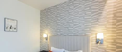 Habitación de hotel con cama, ropa de cama blanca y almohadas rojas. Hay dos mesitas de noche con lámparas, una pared con diseño decorativo y un cuadro enmarcado.
