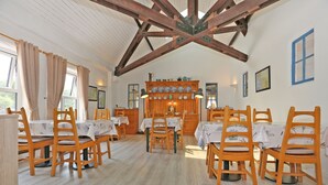 Un comedor con mesas y sillas de madera, techo de madera con vigas a la vista, y una zona de cocina con mostrador de madera y diversos utensilios.