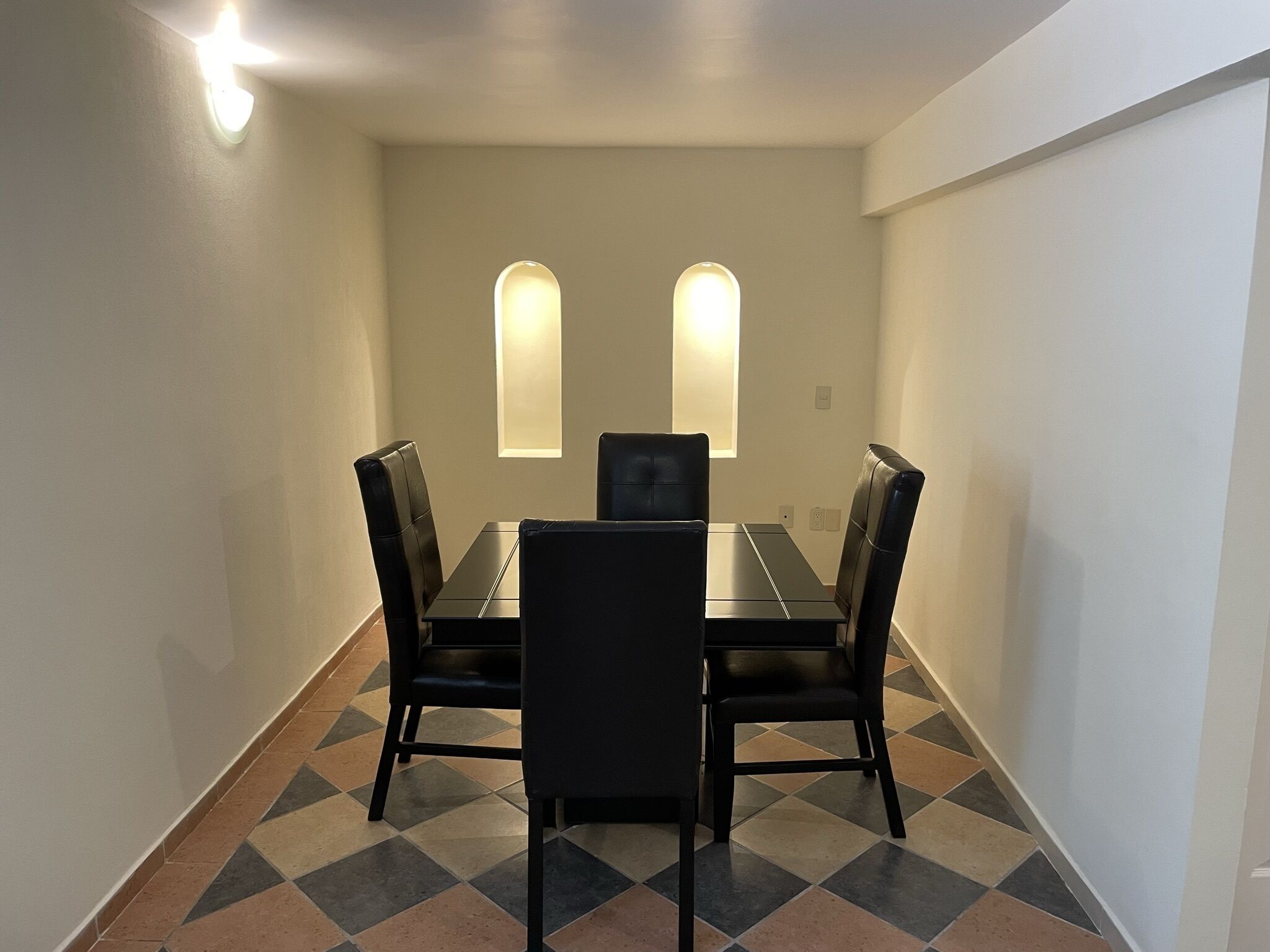 Un comedor con una mesa rectangular y cuatro sillas negras. Dos lámparas están fijadas a la pared sobre la mesa. El suelo presenta un diseño de baldosas de ajedrez.