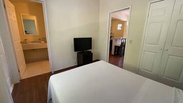 Un dormitorio con cama, televisor, baño con espejo y cocina visible a través de una puerta abierta.