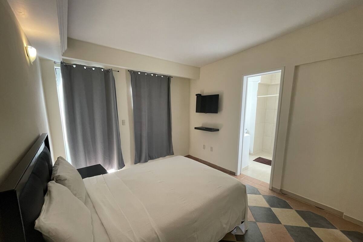 Un dormitorio con cama, cortinas, televisor montado en la pared y un baño visible a través de una puerta abierta.