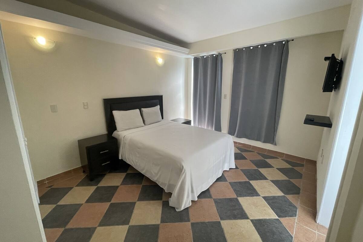 Un dormitorio con cama, mesitas de noche, una ventana con cortinas y suelo de baldosas.