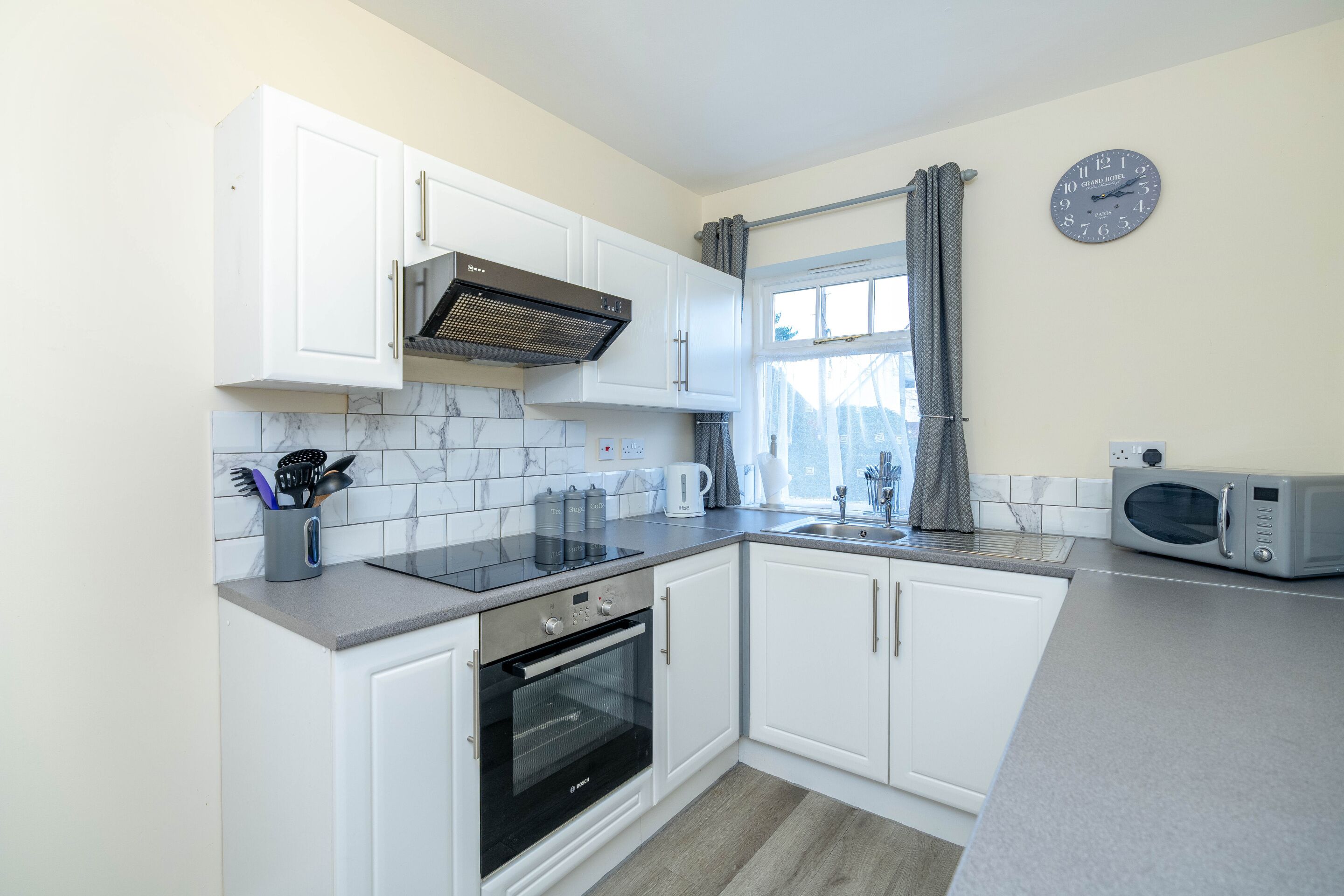 A compact kitchen with white cabinets, a built-in oven, a microwave, and a window with curtains.