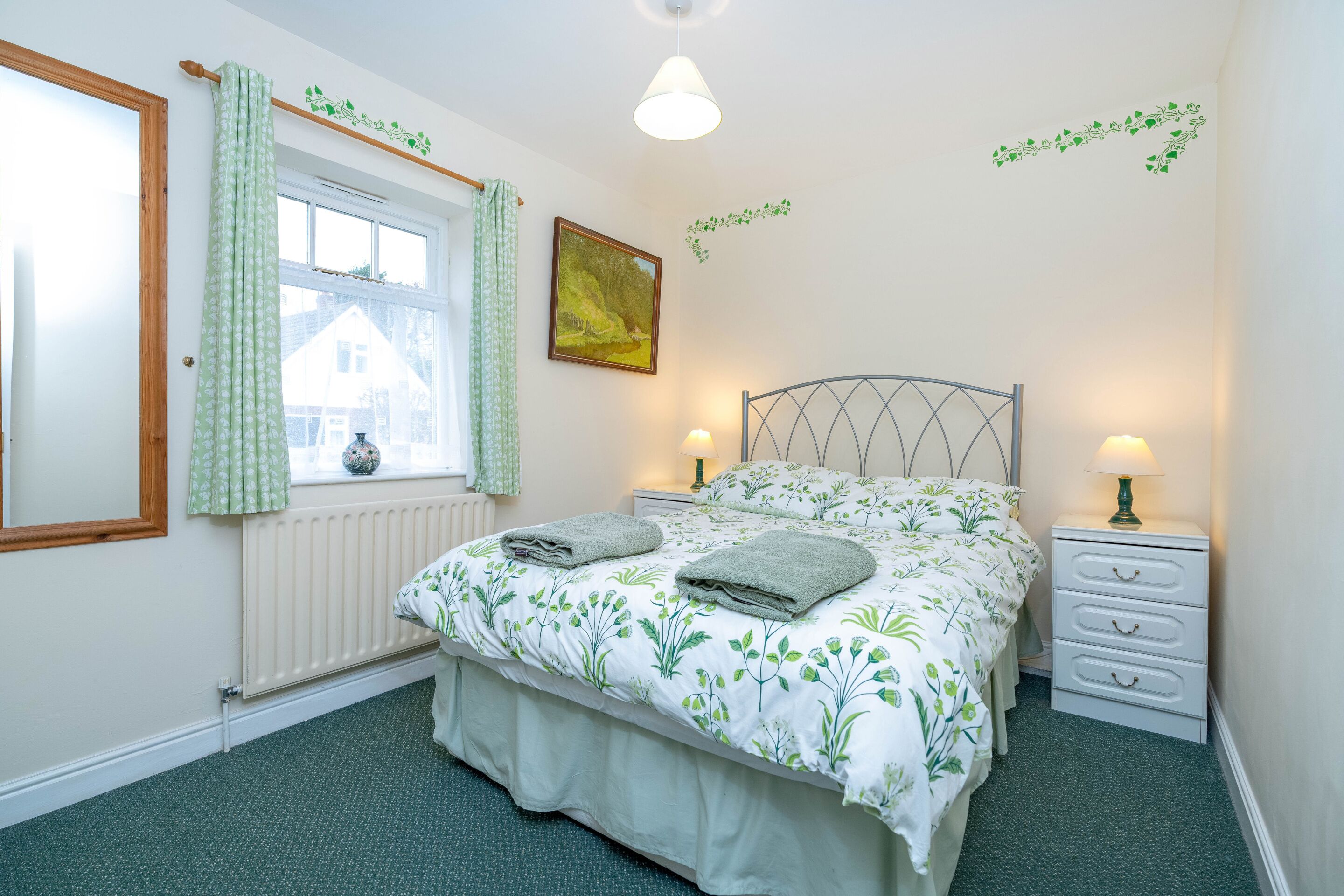 Un dormitorio con una cama, dos mesitas de noche con lámparas, un ventanal con cortinas, un radiador y una pared decorada con un adhesivo de planta.