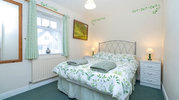 A bedroom with a bed, two bedside tables with lamps, a window with curtains, a radiator, and a wall with a plant decal.