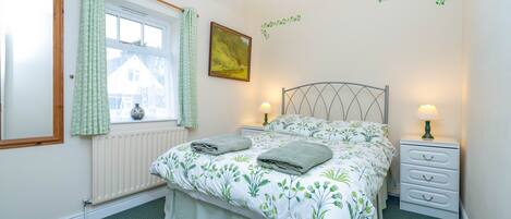 Un dormitorio con una cama, dos mesitas de noche con lámparas, un ventanal con cortinas, un radiador y una pared decorada con un adhesivo de planta.