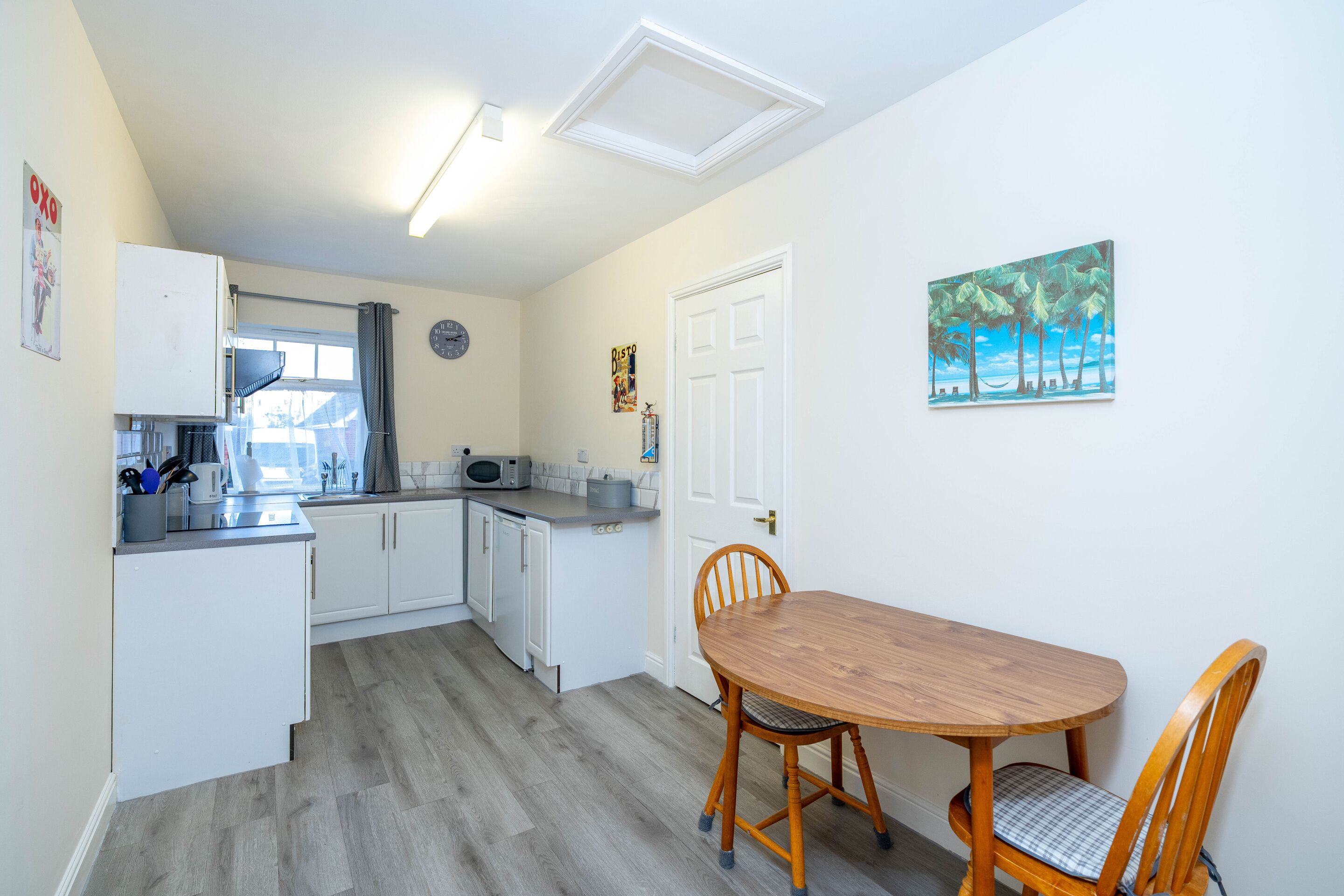 A compact kitchen with white cabinets, a wooden dining table, and a painting of palm trees.