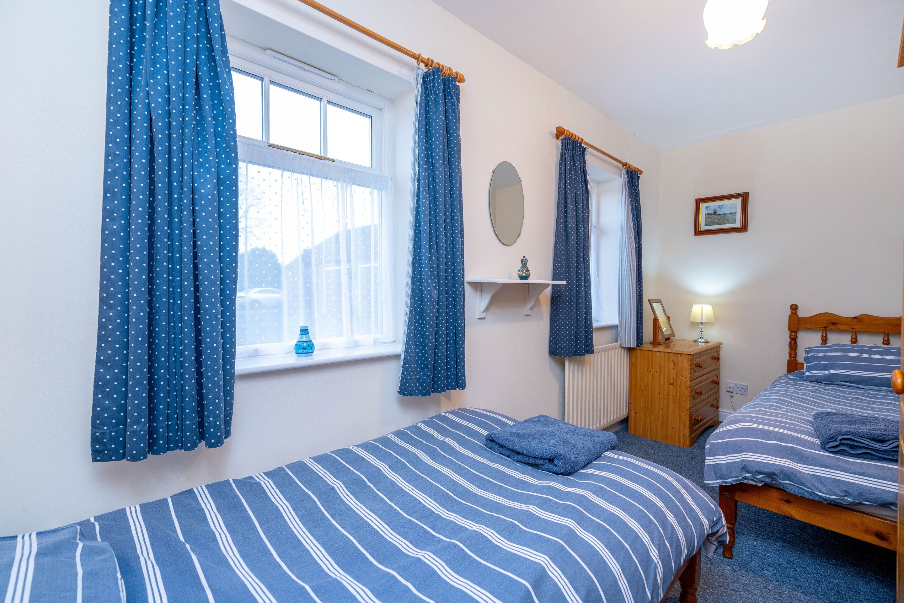 A bedroom with two beds, a window with curtains, a bedside table with a lamp, and a framed picture on the wall.