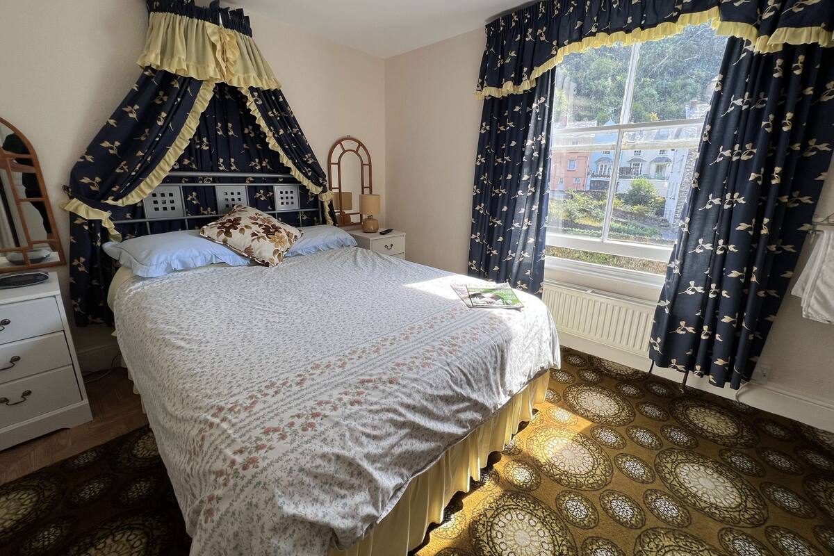 A bedroom with a bed, a nightstand, a lamp, a window with curtains, and a patterned carpet.
