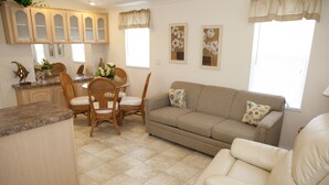 Ein Wohnzimmer mit einem beigefarbenen Sofa, zwei Sesseln, einem Esstisch mit Stühlen und einer Küchenzeile mit Holzschränken und einer Granitarbeitsplatte.