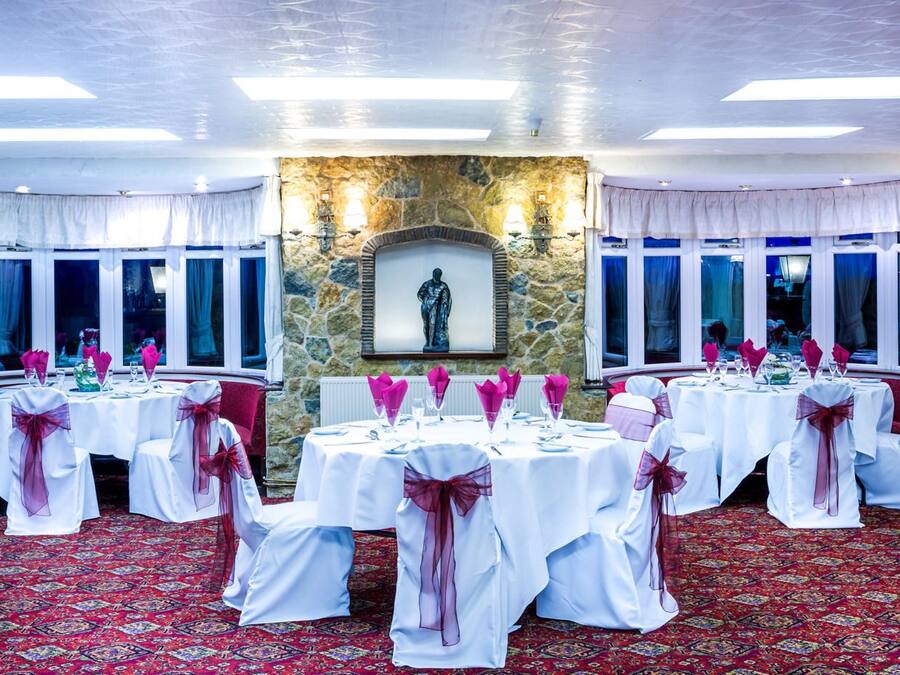 Een feestzaal met ronde tafels, ingericht voor een formeel evenement, met een stenen muur, een standbeeld en grote ramen.