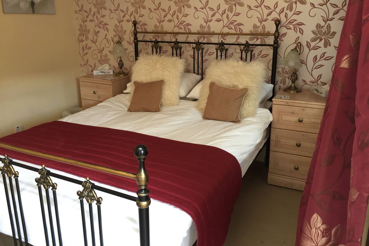A neatly made bed with a red bedspread, two fluffy pillows, and a wooden nightstand with a lamp.
