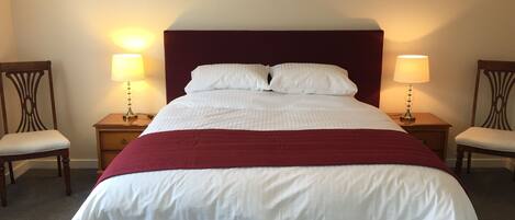 A neatly made bed with white linens and a red accent, flanked by two wooden nightstands with lamps, in a room with plain walls and carpeted floor.