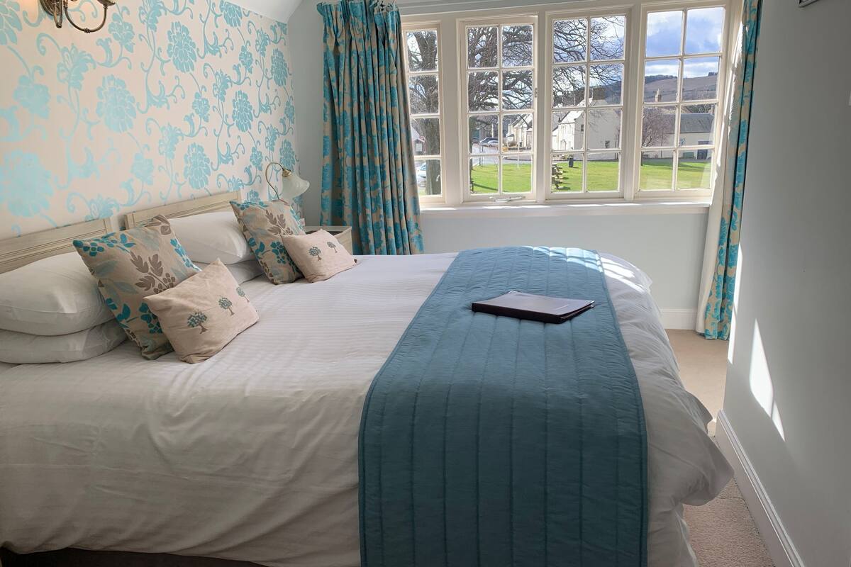 A neatly made bed with a blue quilt and multiple pillows, a window with curtains, and a view of a green landscape.