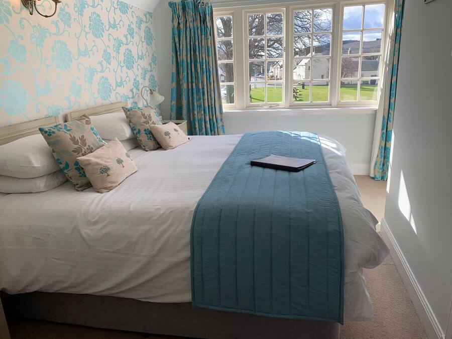 Cama bem arrumada com colcha azul e várias almofadas, janela com cortinas e vista para uma paisagem verde.
