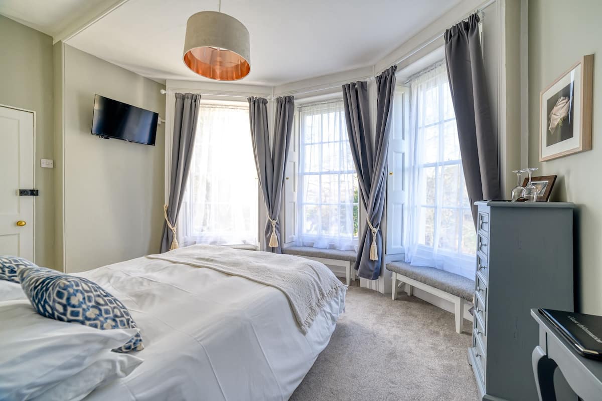 A bedroom with a bed, a desk, a chair, a television, and a window with curtains.