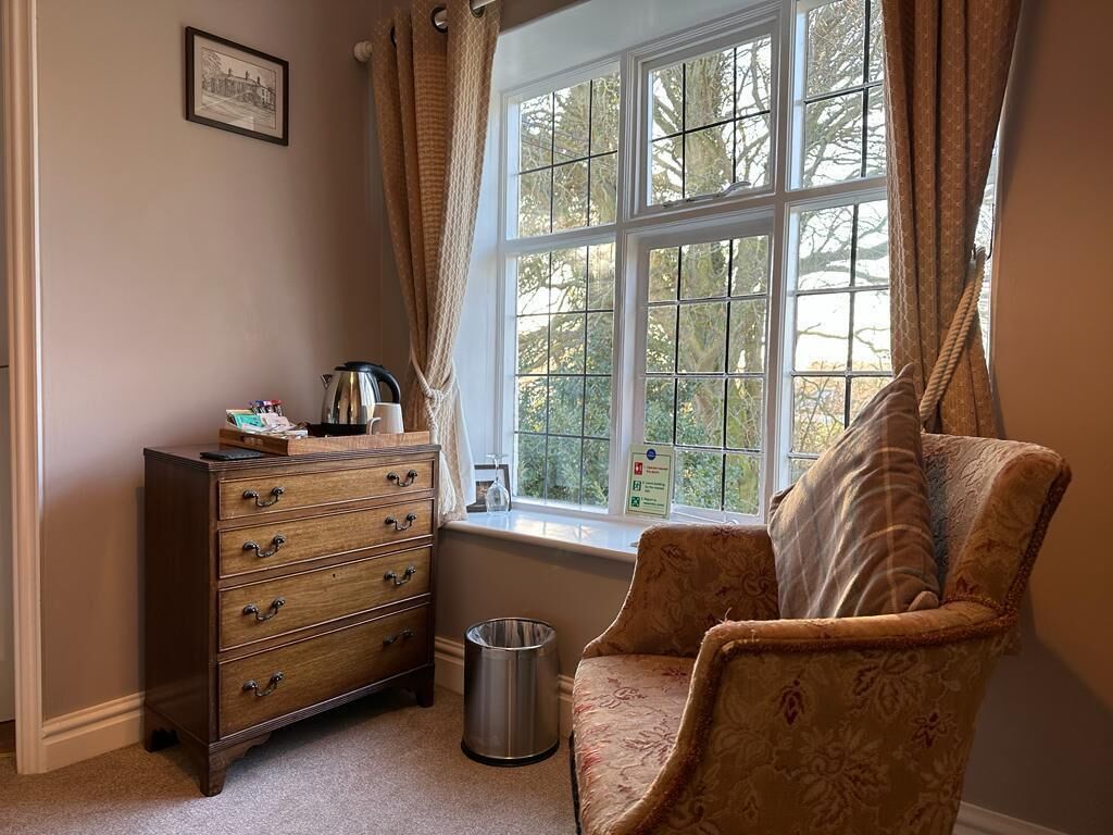 A room with a wooden dresser, a chair, a window with curtains, and a small trash can.