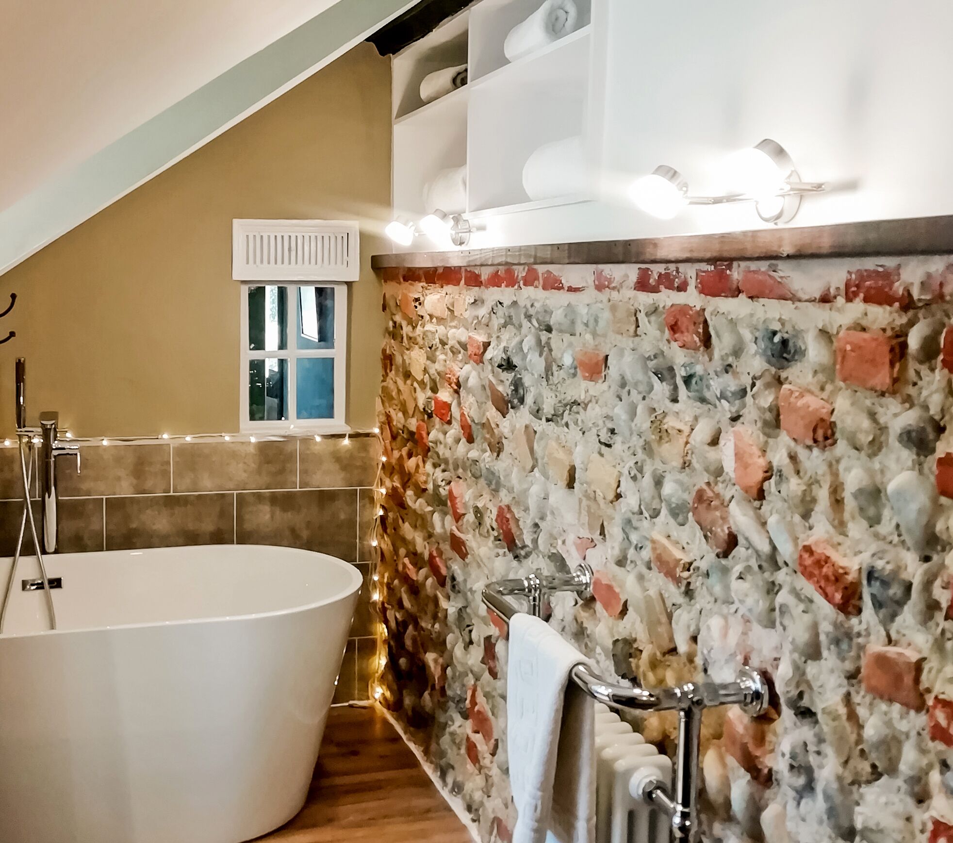 Un baño con una bañera independiente, una pared de mosaico de piedra, una ventana con persiana blanca y un perchero para toallas.
