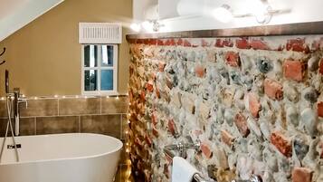 Un baño con una bañera independiente, una pared de mosaico de piedra, una ventana con persiana blanca y un perchero para toallas.