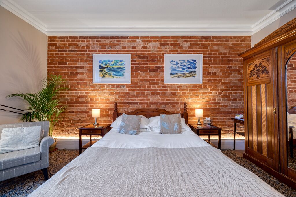 Une chambre d’hôtel avec un mur en briques, un lit avec une literie blanche, deux tables de chevet avec des lampes, une armoire en bois et un fauteuil gris.