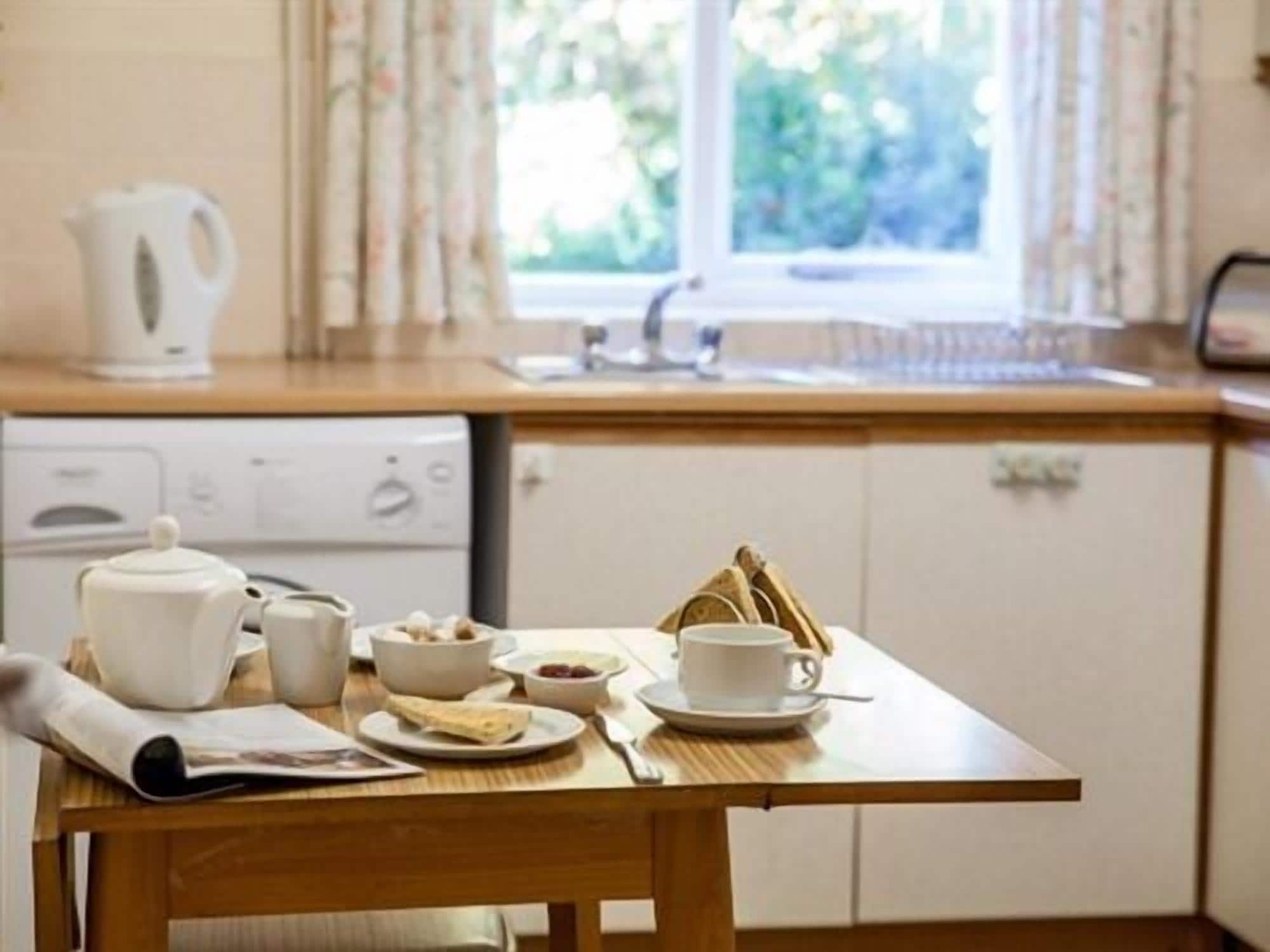 A kitchen with a wooden table set for breakfast, including a kettle, cups, bowls, and a newspaper.