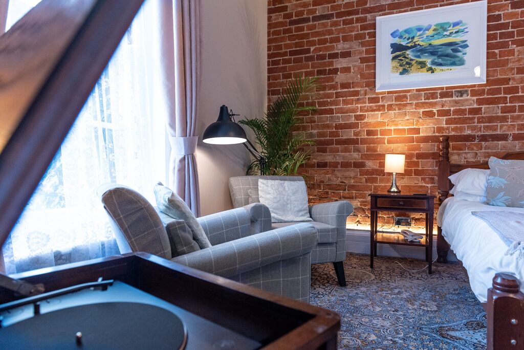 A cozy room with a brick wall, a bed, a desk, a chair, a lamp, and a window with curtains.