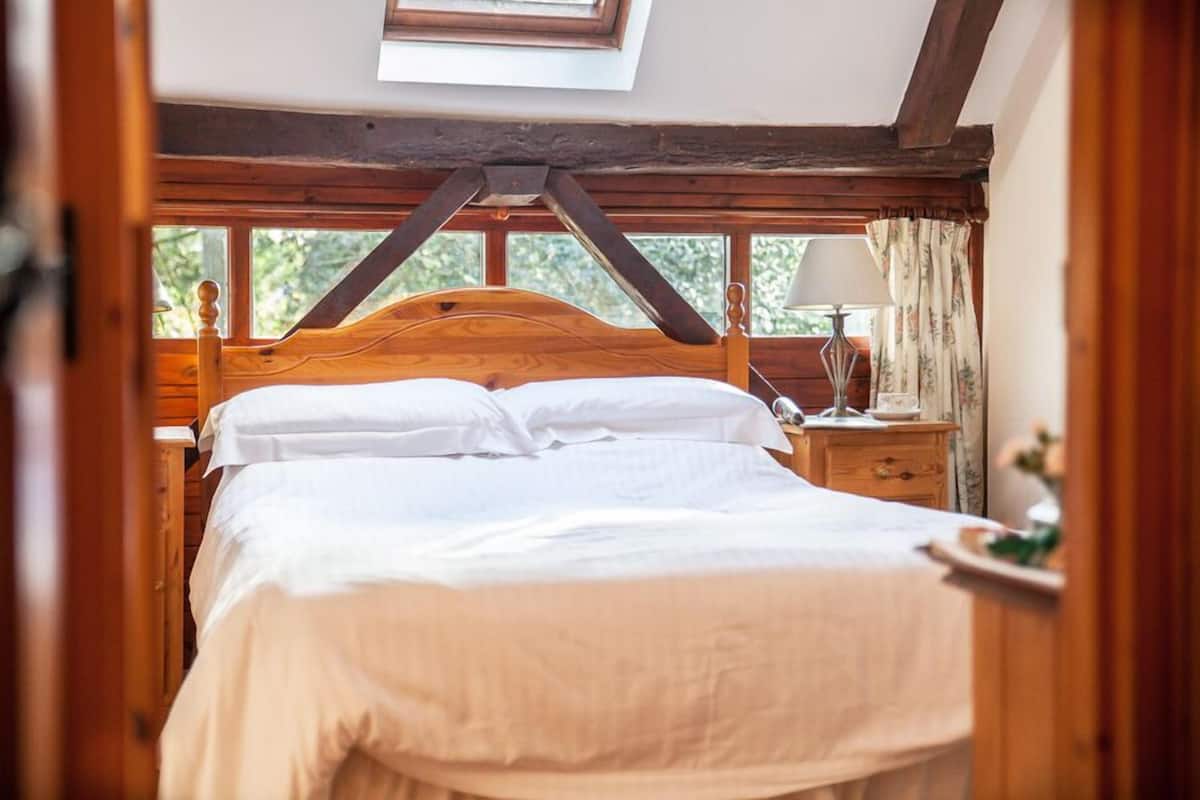 A bedroom with a wooden headboard, a skylight, a bedside table with a lamp, and a window with curtains.