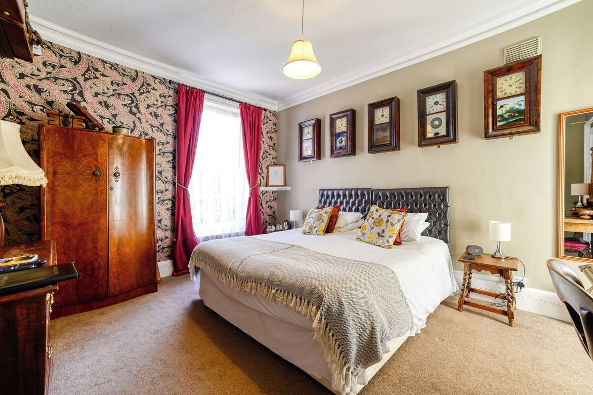 A bedroom with a large bed, a wooden wardrobe, a nightstand with a lamp, and a wall clock.