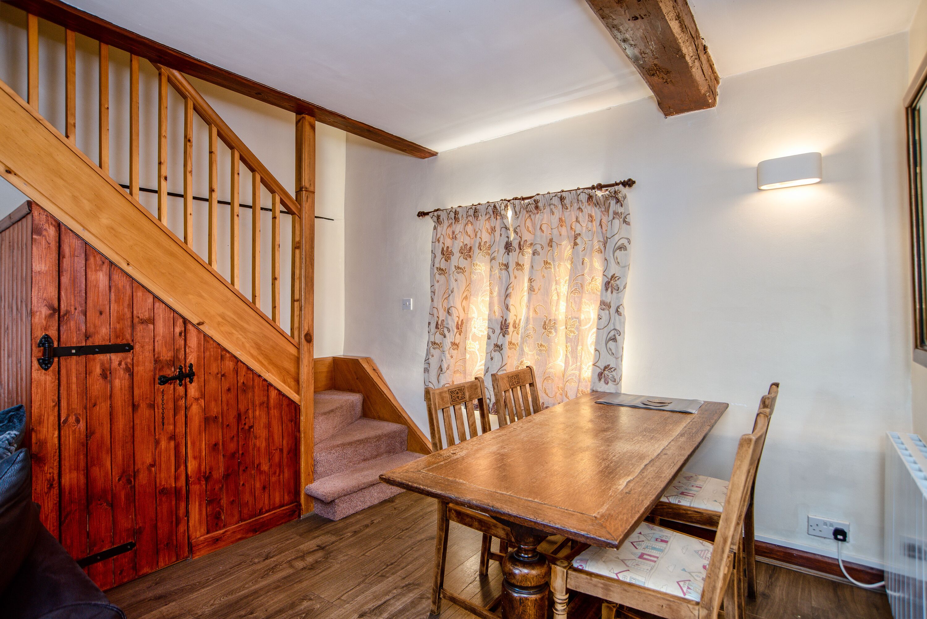 A wooden staircase leading to a room with a dining table and chairs, a window with curtains, and a wall-mounted light fixture.