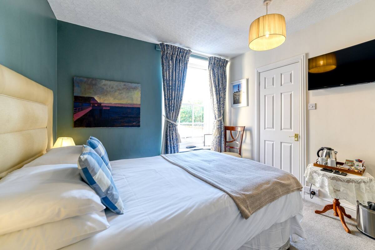 A bedroom with a bed, a bedside table with a kettle, a TV, and a window with curtains.