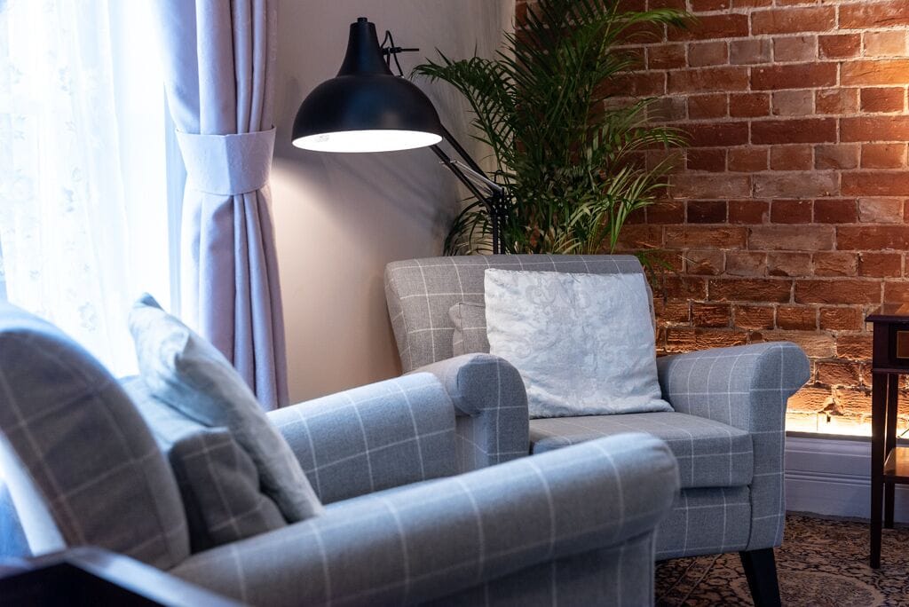 Un salon chaleureux avec un fauteuil gris, un coussin blanc, un lampadaire noir et une plante en pot, le tout adossé à un mur de briques.