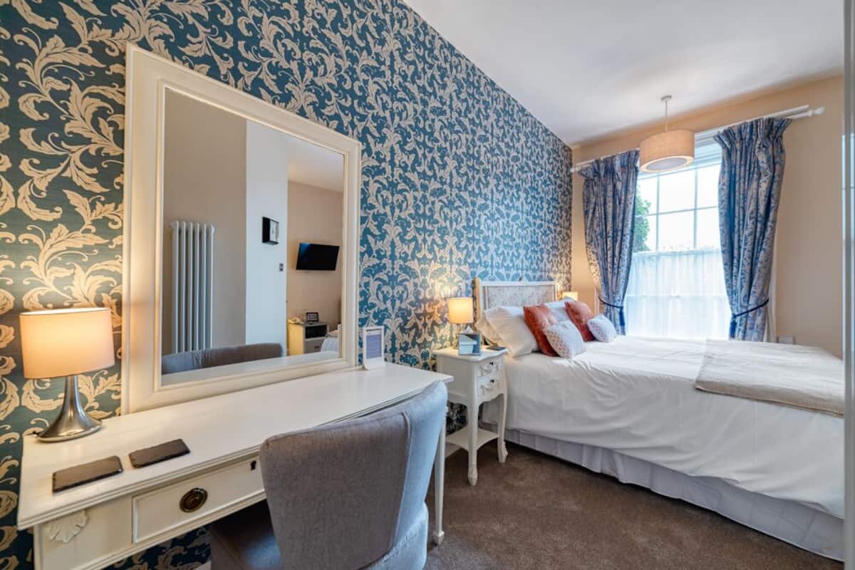 A hotel room with a large mirror, a desk with a lamp, a bed with white bedding, and blue patterned wallpaper.