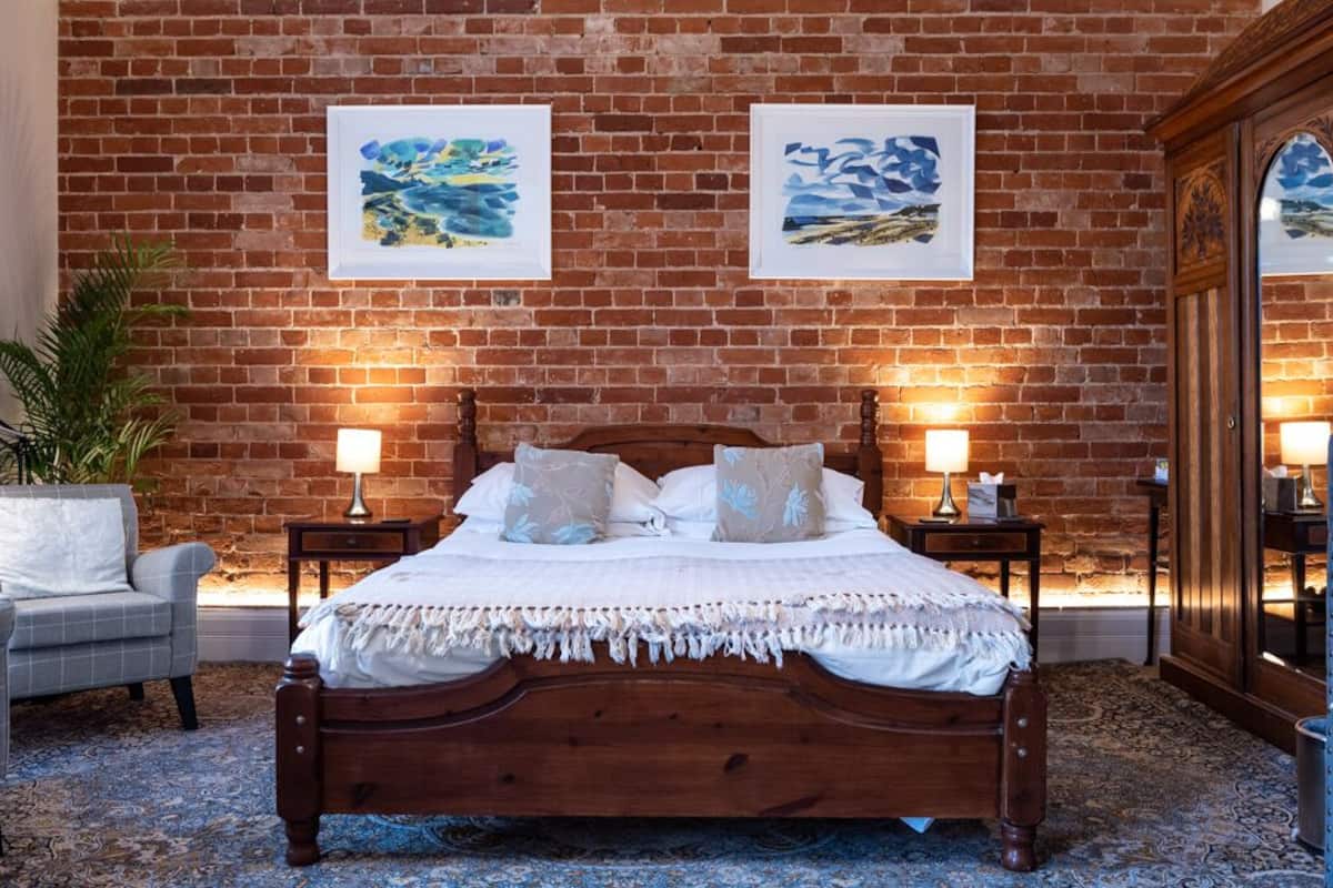 A bedroom with a brick wall, a wooden bed, two bedside tables with lamps, a grey armchair, and a large mirror.
