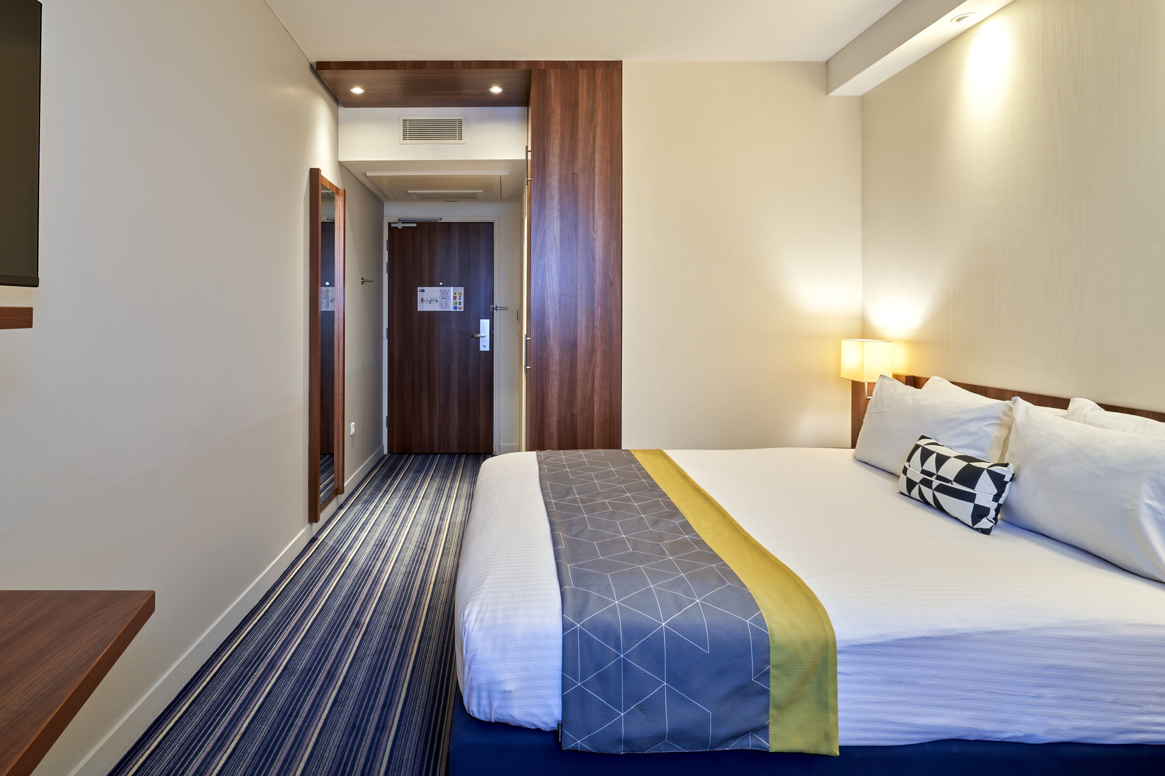 Quarto de hotel com cama, mesa de cabeceira, abajur e porta que dá para outro cômodo.