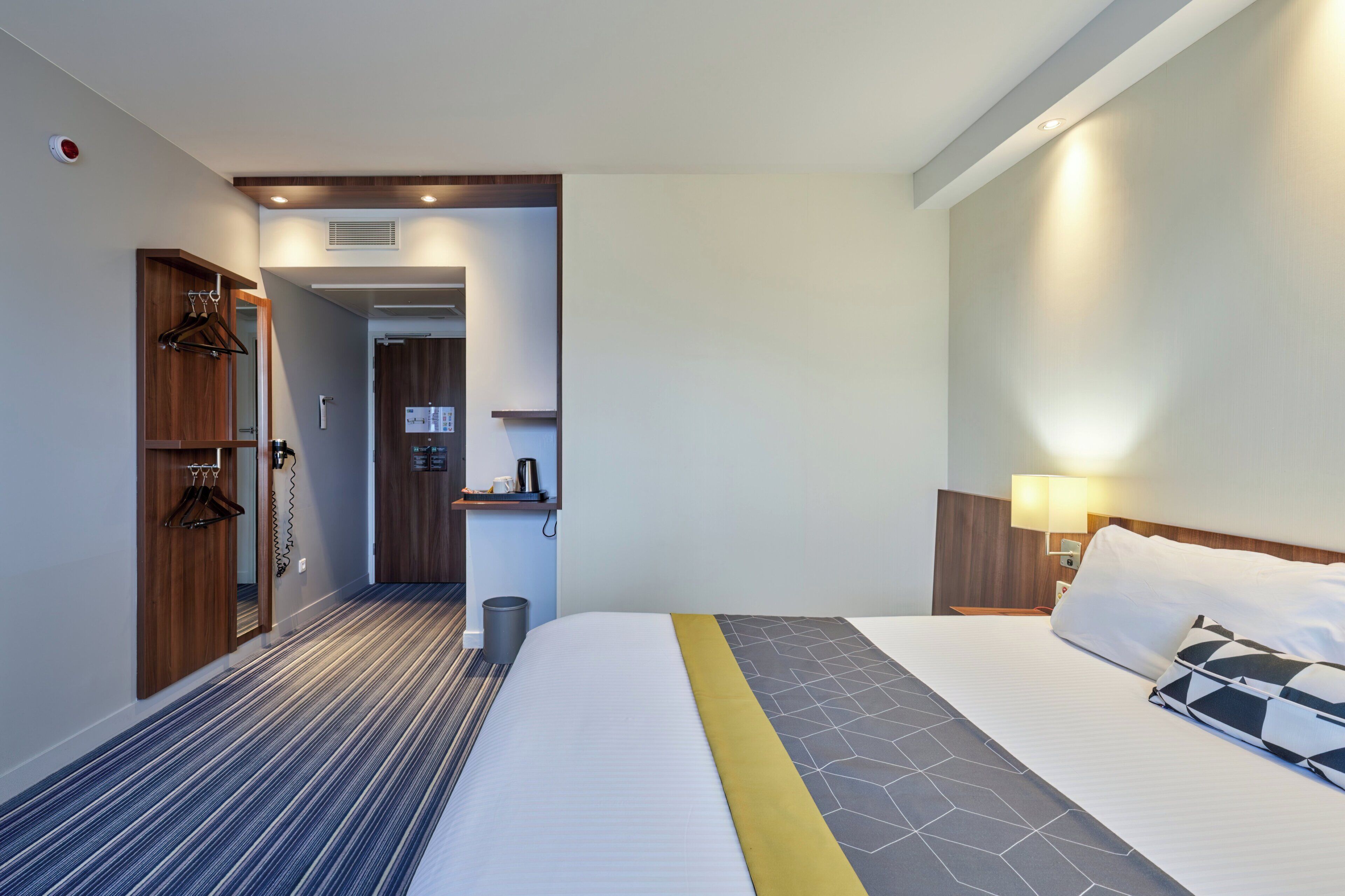 A hotel room with a bed, a bedside table, a lamp, a wardrobe, and a small kitchenette area.