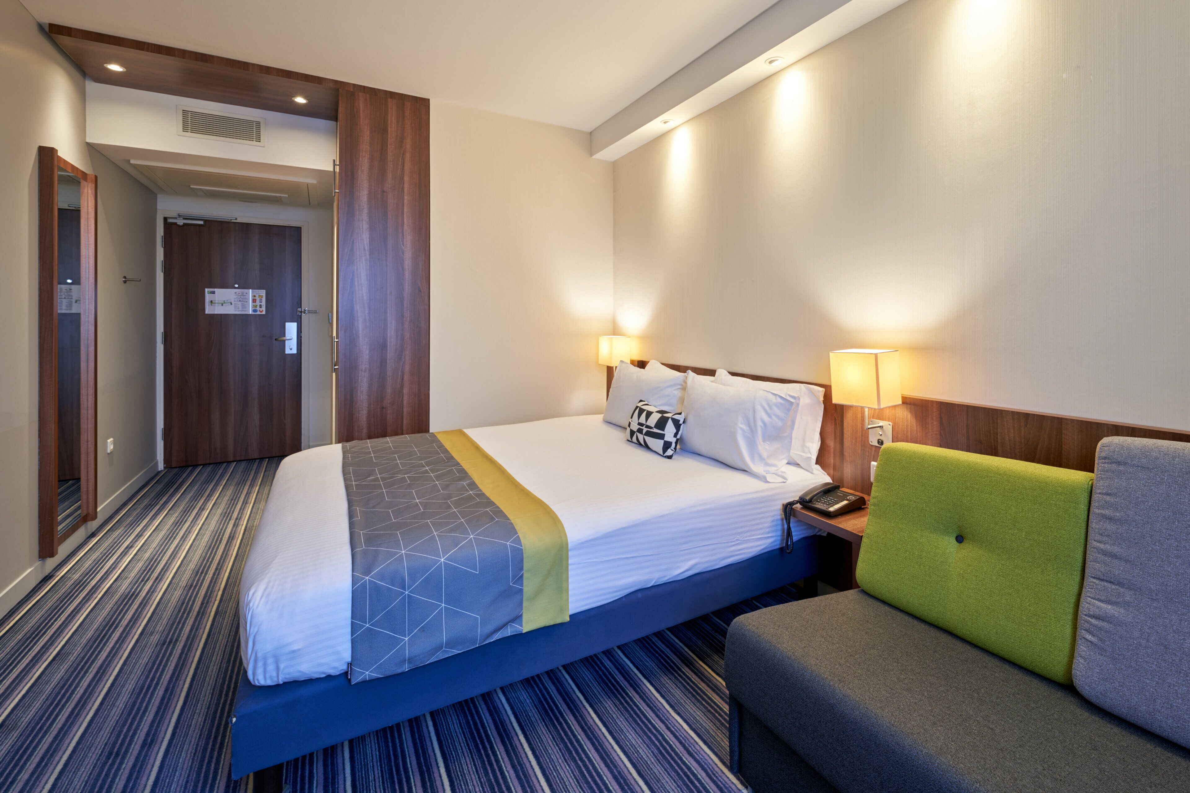 Habitació d'hotel amb un llit, una taula de nit, una làmpada i un sofà.