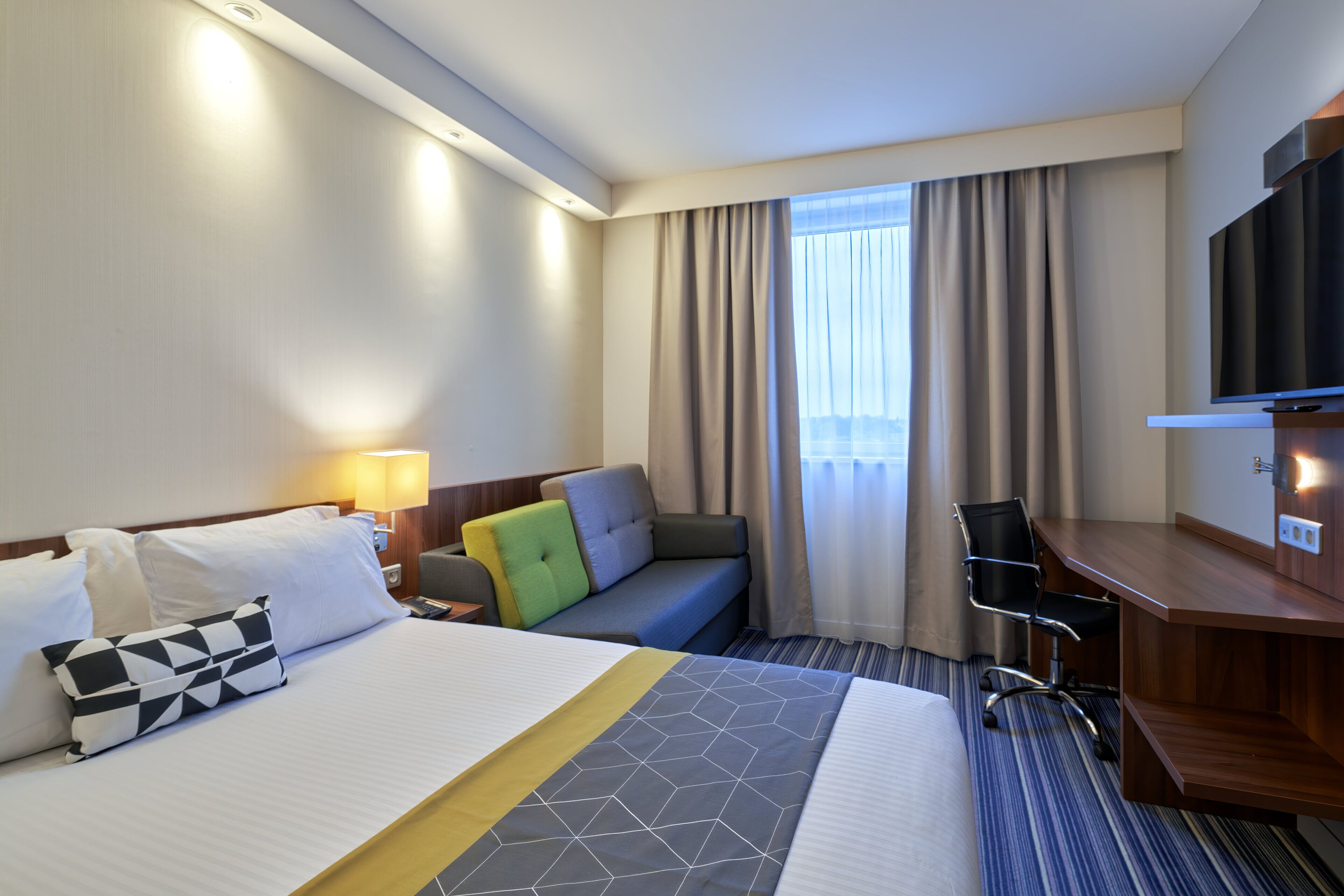 Une chambre d’hôtel avec un grand lit, un bureau, une chaise, une télévision et une fenêtre avec des rideaux.