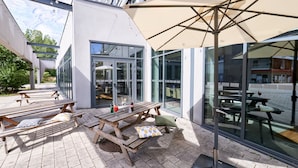 An outdoor patio with wooden tables and benches, a large umbrella, and a glass door leading to an interior space. - Holiday Inn Express Strasbourg - Centre by IHG (Strasbourg)