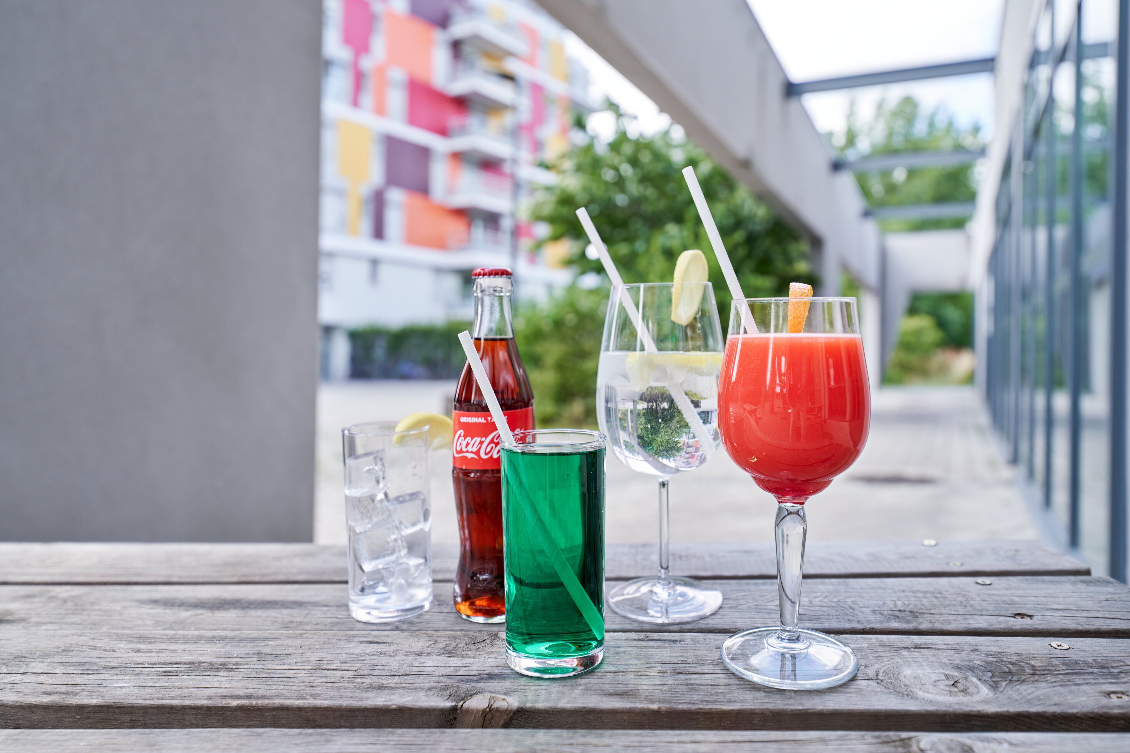 Una mesa de madera con cuatro bebidas: una botella de Coca-Cola, una bebida verde, una bebida roja con una rodaja de limón y una bebida transparente con una pajita.