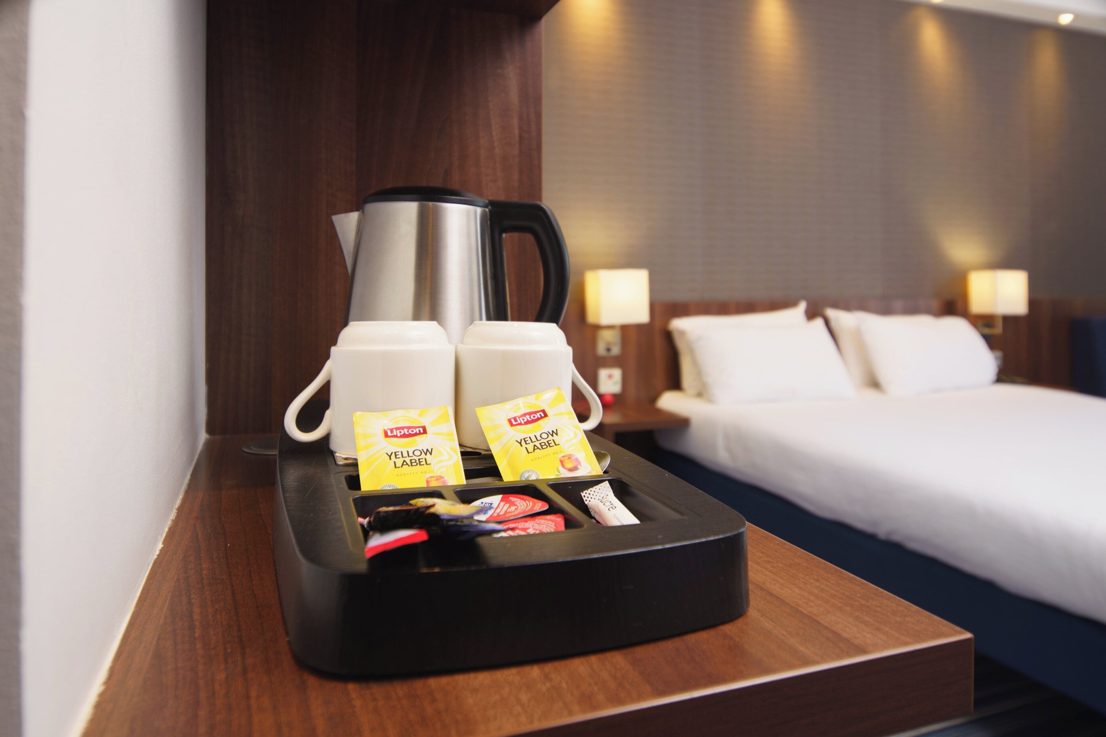 A hotel room with a coffee station featuring a black tray, two white mugs, and Lipton tea packets.