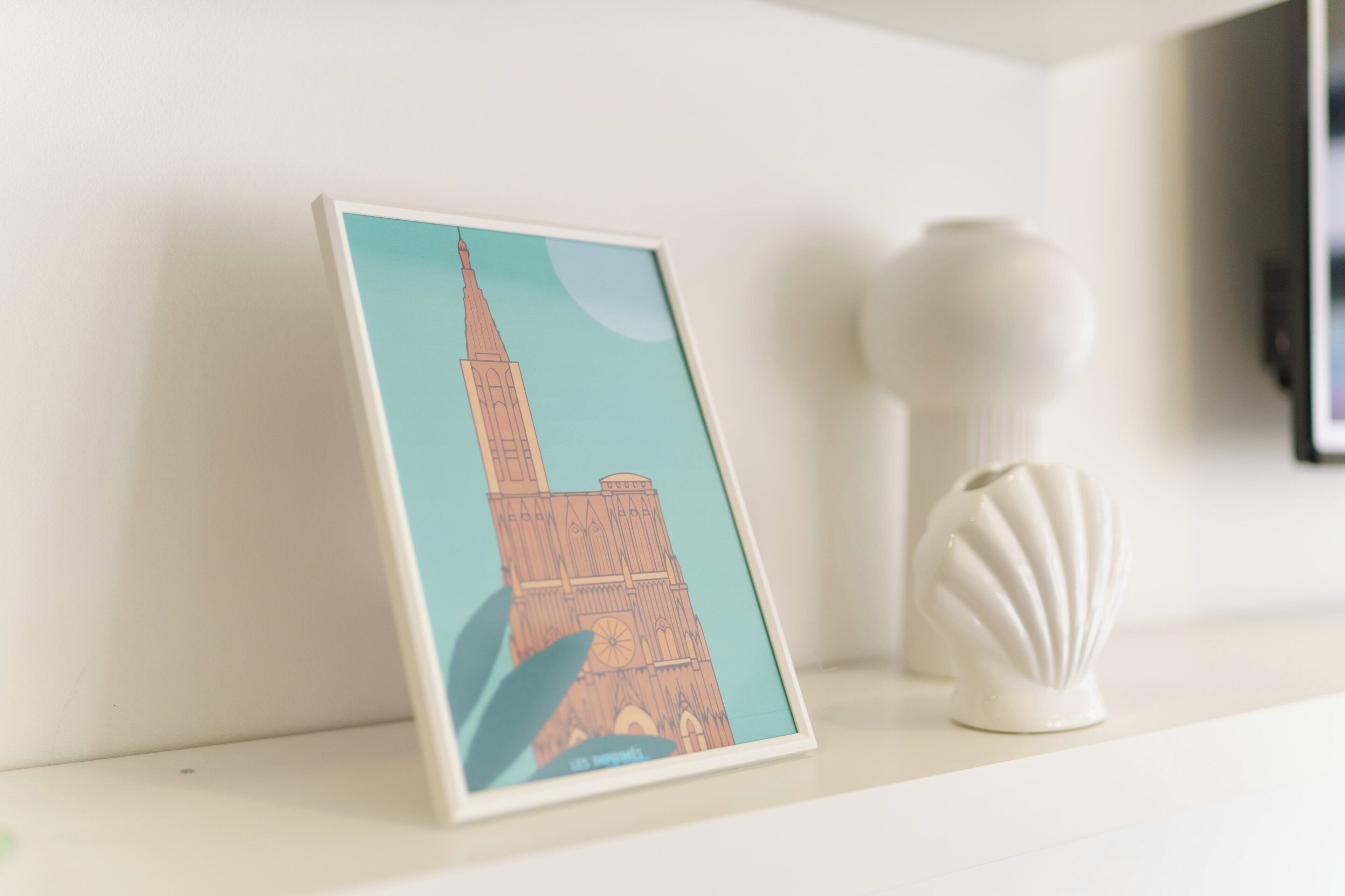 Ein gerahmtes Bild eines Gebäudes mit einem Turm, platziert auf einem Regal neben einer weißen Vase.