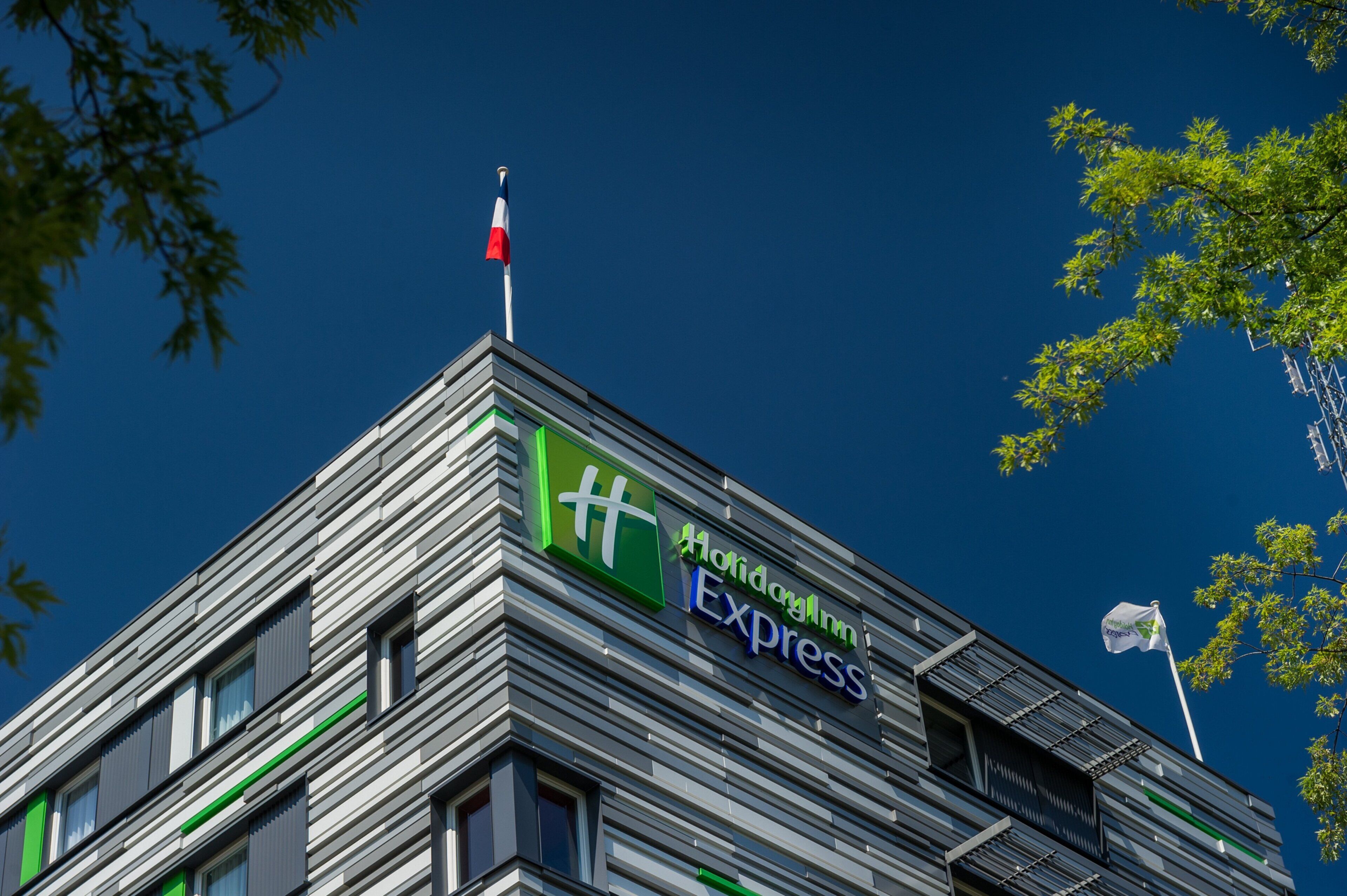 Un edificio moderno con un letrero de Holiday Inn Express, que presenta un asta con una bandera y un cielo azul despejado al fondo.