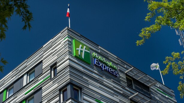 A modern building with a Holiday Inn Express sign, featuring a flagpole with a flag, and a clear blue sky in the background. - Holiday Inn Express Strasbourg - Centre by IHG (Strasbourg)