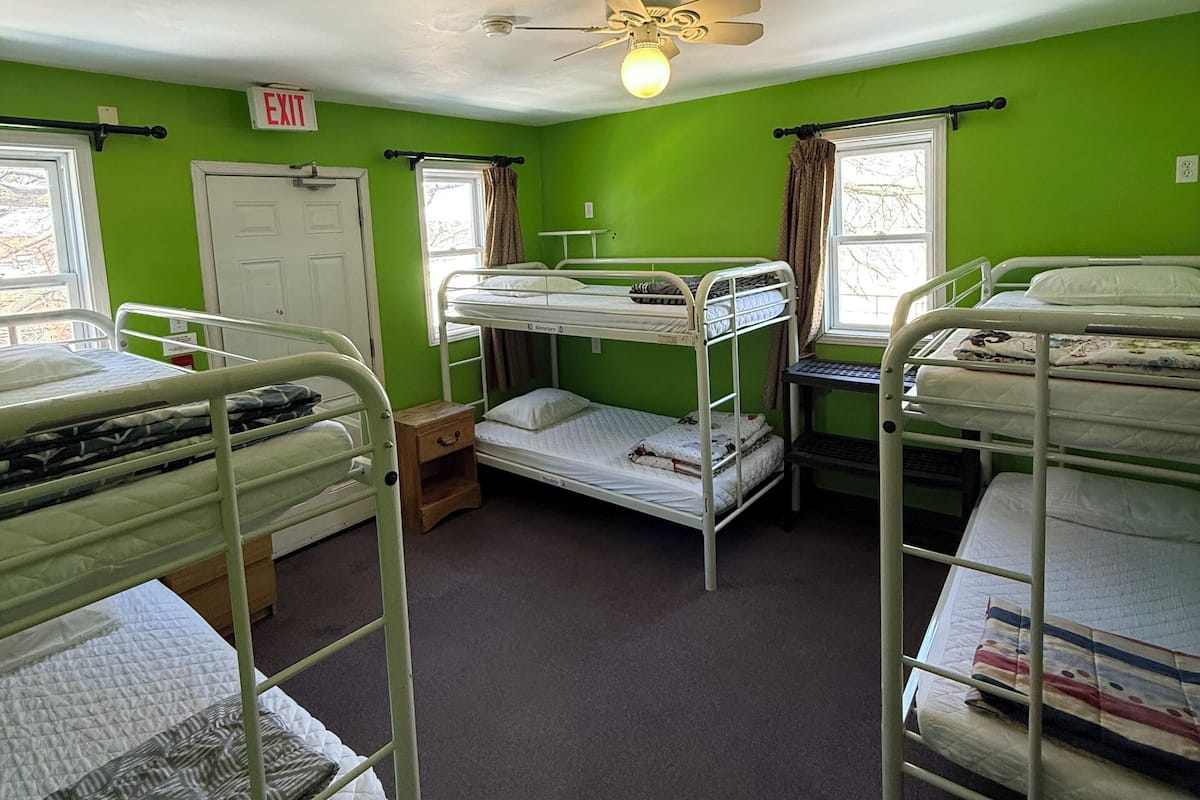 A dormitory room with bunk beds, an exit sign, a ceiling fan, and windows with curtains.