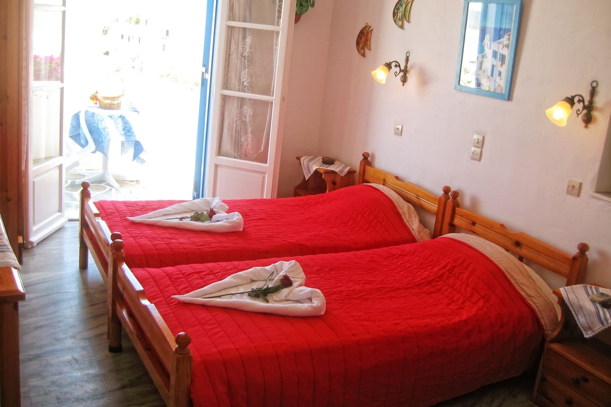 A hotel room with two beds, each with a red bedspread and a towel with a rose. There is a window with a view of the outside, a wall with decorative items, and a small table with a lamp.