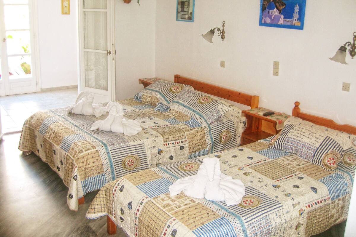 A room with two beds, each with a quilted bedspread and a wooden headboard. There is a small bedside table with a lamp on one side and a wall-mounted lamp on the other. A potted plant hangs on the wall, and there are two framed pictures.